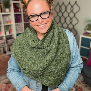 May include: A green knitted cowl scarf is worn over a denim jacket. The cowl is a solid olive green color and appears to be hand-knitted. The person in the image is wearing black-framed glasses and smiling.