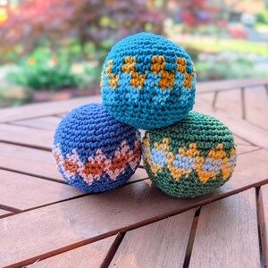 Pyramid of three crochet hacky sacks in blue, teal and green with zigzag stripes on a wooden table.