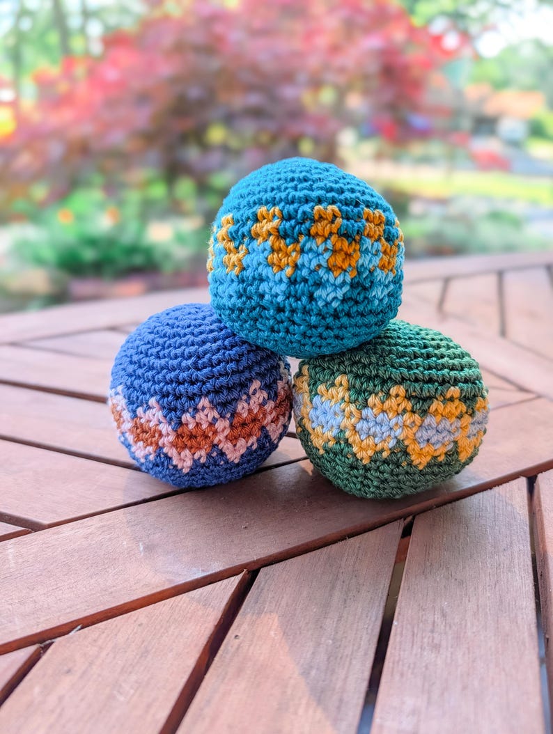 Pyramid of three crochet hacky sacks in blue, teal and green with zigzag stripes on a wooden table.