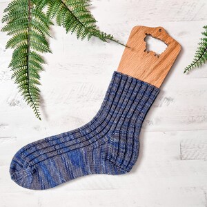 A single blue and gray ribbed knit sock is displayed on a wooden sock blocker, placed on a white wooden surface with two green fern leaves nearby—perfect as a handmade gift or for pairing with your favorite vintage clothes.