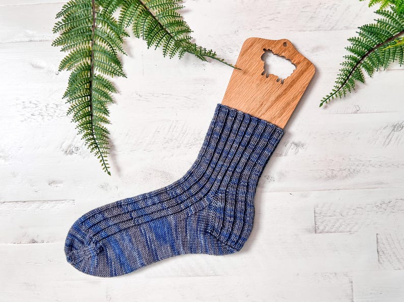 A single blue and gray ribbed knit sock is displayed on a wooden sock blocker, placed on a white wooden surface with two green fern leaves nearby—perfect as a handmade gift or for pairing with your favorite vintage clothes.