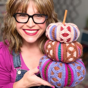 May include: A stack of three crocheted pumpkins featuring intricate geometric patterns in shades of pink, brown, purple, teal, and green. The top pumpkin is topped with a brown cinnamon stick stem, and each pumpkin has a textured, ribbed surface.
