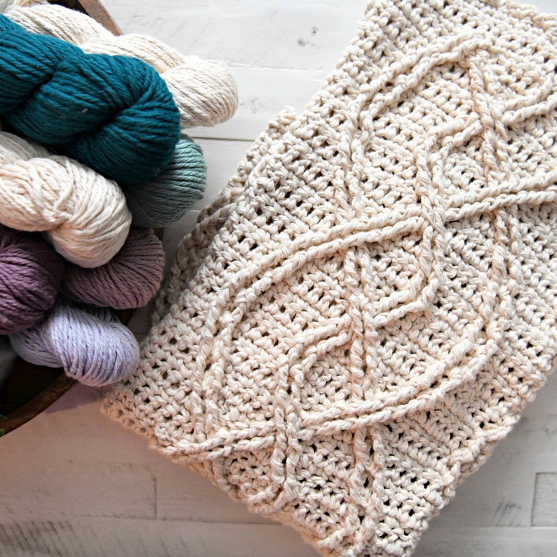 A crochet project in progress with a beautiful textured pattern, surrounded by skeins of colorful yarn and crochet tools, evoking a sense of creativity and craftsmanship for a unique handmade gift. Inishmore Crochet Cable Blanket - Marly Bird