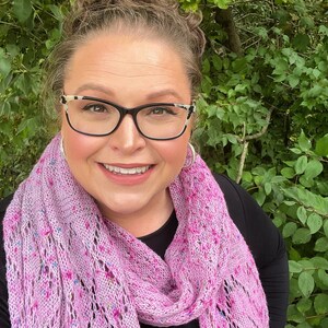 Knit and Crochet Designer Marly Bird is modeling her Diamonds and Pearls Knit Shawl made in a speckled yarn with a pink base. She is standing outdoors surrounded by greenery.