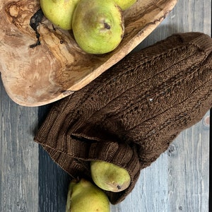 A rustic still life with ripe green pears on a textured wooden bowl and a knitted brown market bag, all set on a weathered wooden surface alongside unique jewelry.