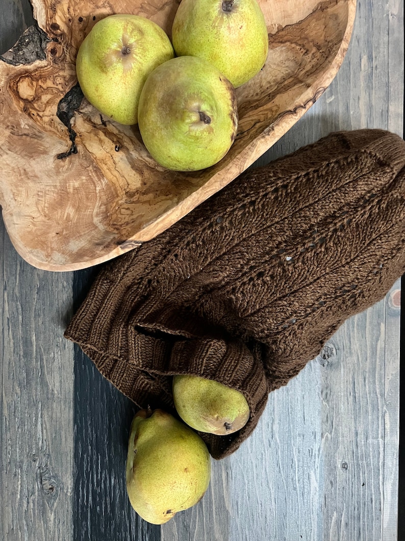 A rustic still life with ripe green pears on a textured wooden bowl and a knitted brown market bag, all set on a weathered wooden surface alongside unique jewelry.