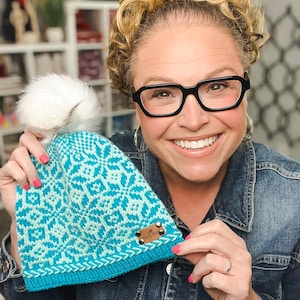 Peut inclure: Un bonnet en tricot avec un motif géométrique de flocons de neige bleu sarcelle et bleu clair, un pompon en fausse fourrure blanche sur le dessus et une étiquette rectangulaire en cuir marron avec le texte Marly Bird fixé par quatre rivets.