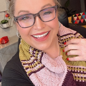 three color knit bandana cowl worn around the neck of Marly Bird. The bind off edge is seamed to the bottom ridge edge near the cast on to form a point in front when wearing it. The cowl is made in light pink, wine purple, and a golden wheat color.