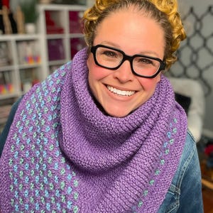 May include: A person wearing a knitted purple scarf. The scarf features a textured pattern with blue and green accents on one side. The person is smiling and wearing black glasses.