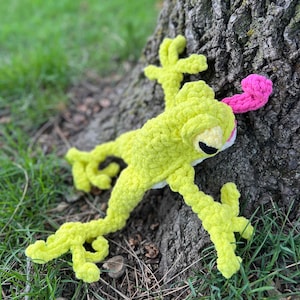 May include: A crocheted green frog toy with a pink tongue sticking out. The frog is sitting on a tree trunk with green grass in the background.