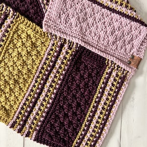 three color knit bandana cowl flat on a wood surface to show the 3d seam. This seam and the construction of the cowl form a point in front when wearing it. The cowl is made in light pink, wine purple, and a golden wheat color.