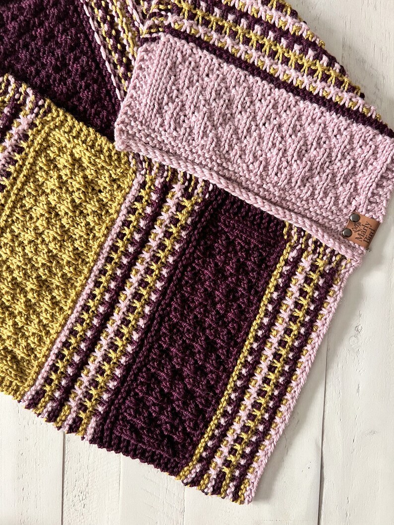 three color knit bandana cowl flat on a wood surface to show the 3d seam. This seam and the construction of the cowl form a point in front when wearing it. The cowl is made in light pink, wine purple, and a golden wheat color.