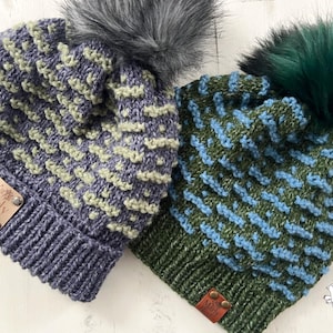 May include: Two knitted hats with pom poms. The hats are made with a textured stitch and have a contrasting color stripe. One hat is blue and green, the other is gray and green. Both hats have a leather tag with the words "Marly Bird" on them.