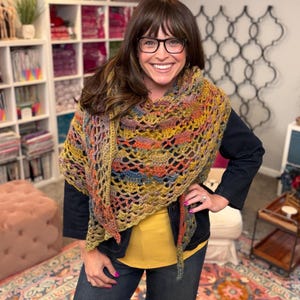 May include: A triangular crochet shawl featuring a variegated yarn pattern with blended tones of mustard yellow, burnt orange, deep blue, and earthy brown. The accessory has an open, lacy stitch design with a scalloped edge, draped over a dark jacket.