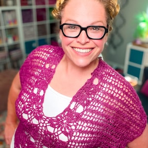 May include: A magenta crochet top with a floral design and a deep V-neck. The top is worn over a white undershirt. The crochet work has an open, airy pattern. The top is a vibrant shade of pink.
