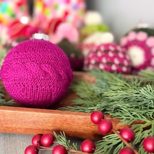 May include: A spherical Christmas ornament covered in a vibrant magenta knitted fabric featuring a horizontal cable knit pattern around the center. The ornament has a silver metal cap at the top and is displayed alongside green pine needles.