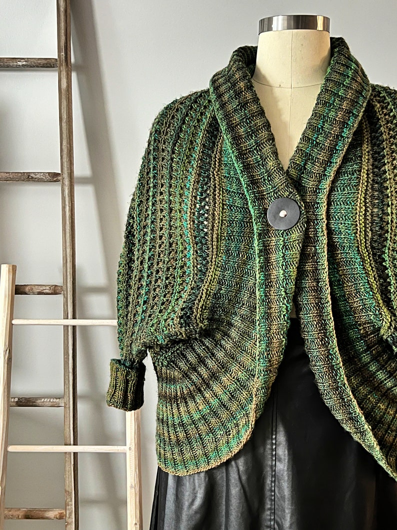 An elegantly draped, hand-knit cocoon cardigan displayed on a mannequin, showcasing rich hues of green with a subtle variegated pattern, chunky ribbed collar, and a large decorative button at the front. Marly Bird Know Your Worth Knit Cocoon Cardigan