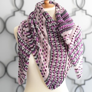 May include: A knitted shawl in shades of purple, gray, and white, displayed on a mannequin. The shawl features a geometric pattern with a triangular shape, perfect for wrapping around the shoulders. The design includes various textures and color blocks.