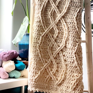 The cozy Inishmore Crochet Cabled Blanket is displayed on a wood ladder. this hand-crocheted blanket with intricate cable patterns is in front of a stack of colorful bulky cotton yarn hanks and a potted plant in the background. Marly Bird