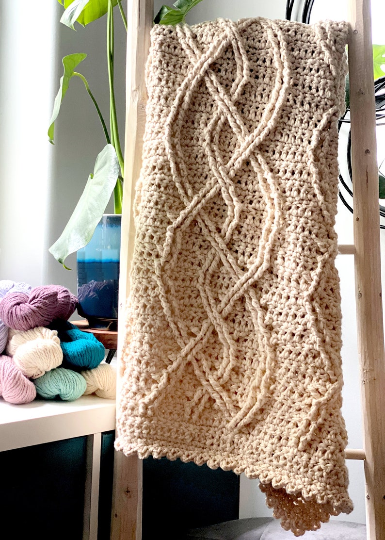 The cozy Inishmore Crochet Cabled Blanket is displayed on a wood ladder. this hand-crocheted blanket with intricate cable patterns is in front of a stack of colorful bulky cotton yarn hanks and a potted plant in the background. Marly Bird
