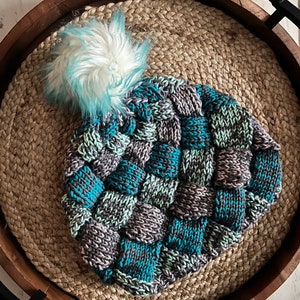 Puede incluir: Un gorro de punto azul, gris y blanco con un pompón blanco esponjoso. El gorro está hecho con un hilo grueso y tiene un patrón tejido único.