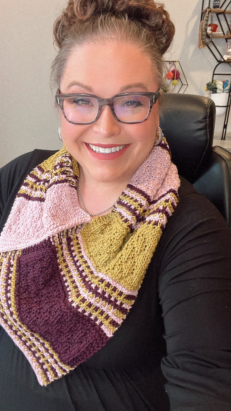 three color knit bandana cowl worn around the neck of Marly Bird. The bind off edge is seamed to the bottom ridge edge near the cast on to form a point in front when wearing it. The cowl is made in light pink, wine purple, and a golden wheat color.