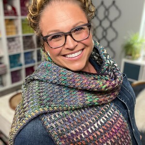 May include: A colorful, hand-knitted scarf with a textured pattern. The scarf features a mix of colors including purple, green, orange, and white. The scarf is wrapped around the neck and shoulders.