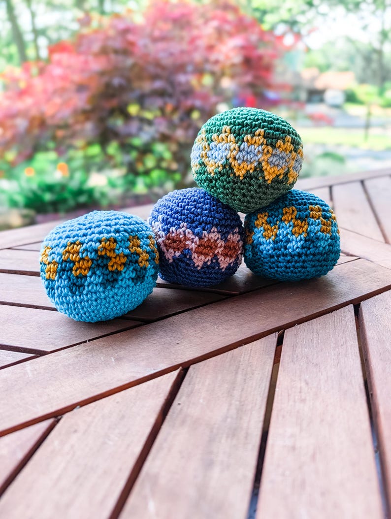 Three crochet hacky sacks in blue, navy and green with orange and gold zigzag colorwork stacked on a wooden outdoor table.