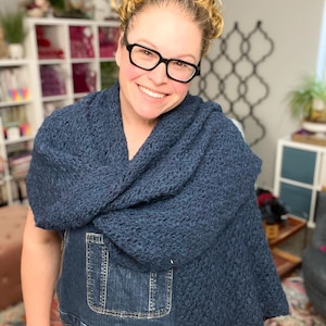 May include: A woman wearing a dark blue, textured, crocheted shawl. The shawl is draped over the shoulders and chest. The woman is wearing black glasses and denim overalls. The background includes shelves and decorative items.