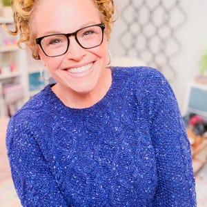 May include: A long-sleeved, crew-neck sweater in a vibrant royal blue color. The knit fabric features a subtle cable-knit pattern throughout and is speckled with tiny flecks of lighter blue, purple, and white, creating a textured appearance.
