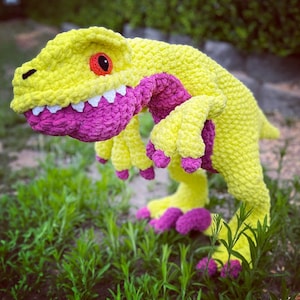 Puede incluir: Un juguete de dinosaurio de crochet con cuerpo amarillo y detalles rosas. El dinosaurio está de pie sobre sus patas traseras con la cola enroscada.