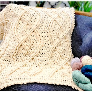 A close-up of a cream-colored crochet blanket with intricate cable patterns, resting against a chair, with a blurred background suggesting a cozy interior space. skeins of yarn in muted colors are visible in the lower right corner - Marly Bird