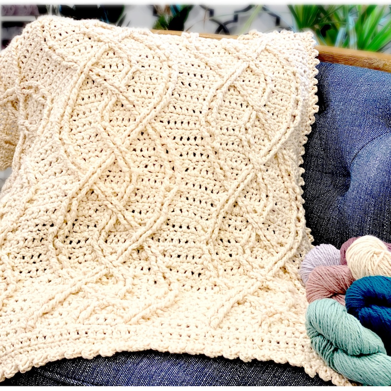 A close-up of a cream-colored crochet blanket with intricate cable patterns, resting against a chair, with a blurred background suggesting a cozy interior space. skeins of yarn in muted colors are visible in the lower right corner - Marly Bird