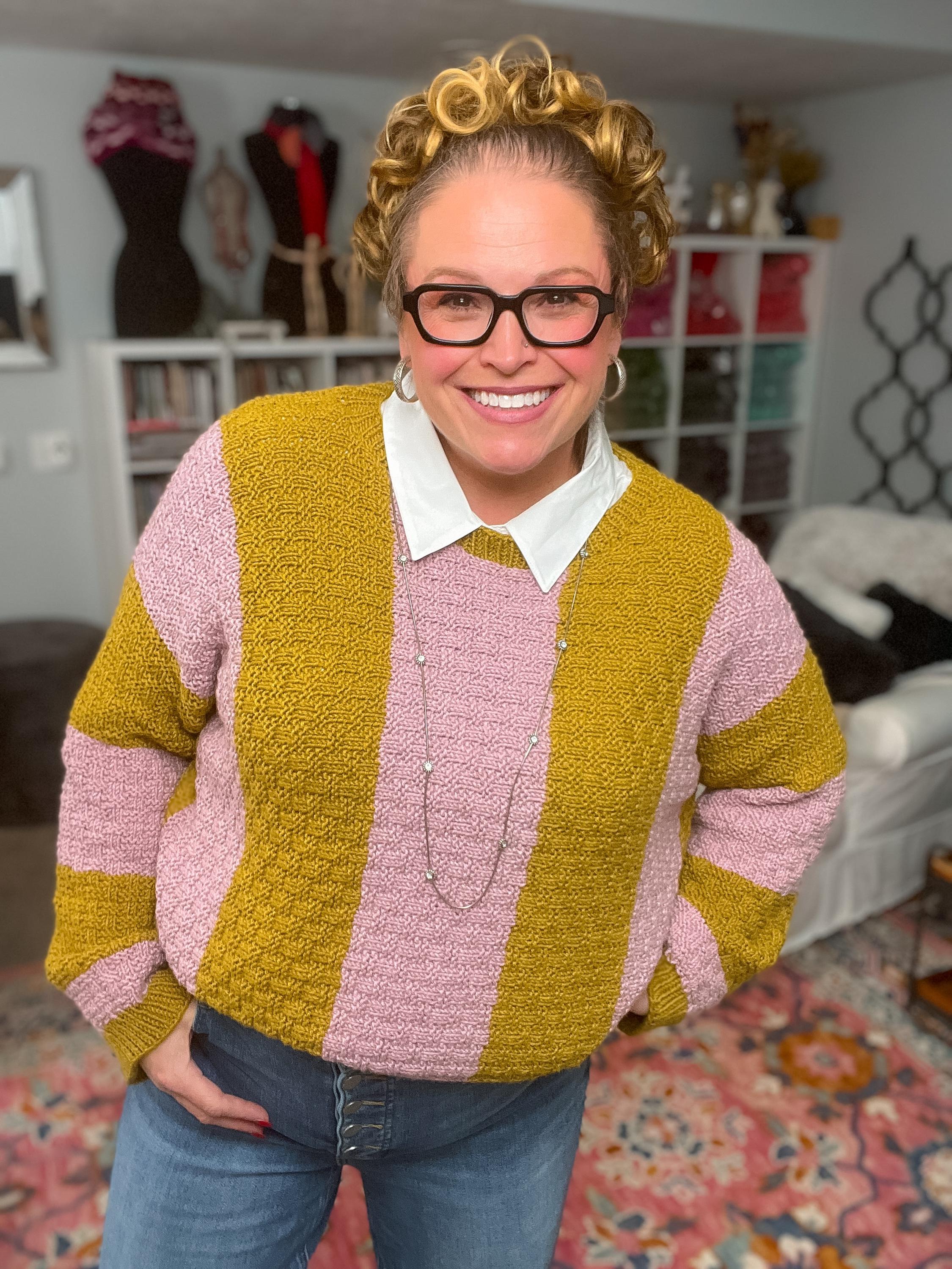 Könnte beinhalten: Ein gestreifter Pullover in Senfgelb und Rosa. Der Pullover hat ein strukturiertes Strickmuster und wird über einem weißen Hemdkragen getragen. Die Person trägt eine blaue Jeans und eine silberne Halskette. Der Hintergrund umfasst ein Bücherregal und einen gemusterten Teppich.