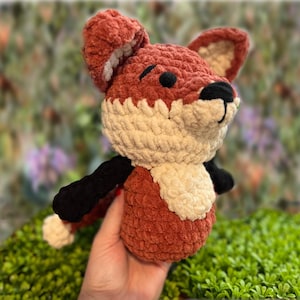 May include: A handmade plush fox toy in shades of rust, cream, black, and tan. The fox has a fluffy texture, with a white chest and inner ears. The fox's arms and nose are black. The toy is held in a hand.