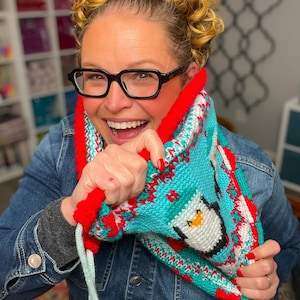 Könnte beinhalten: Ein gestrickter Cowl-Schal mit Pinguin-Design. Der Schal ist hauptsächlich türkis mit roten, weißen und schwarzen Akzenten. Der Pinguin hat einen weißen Körper, schwarze Flügel und einen gelben Schnabel. Der Cowl hat einen roten Rand und hellblaue Kordelzüge.