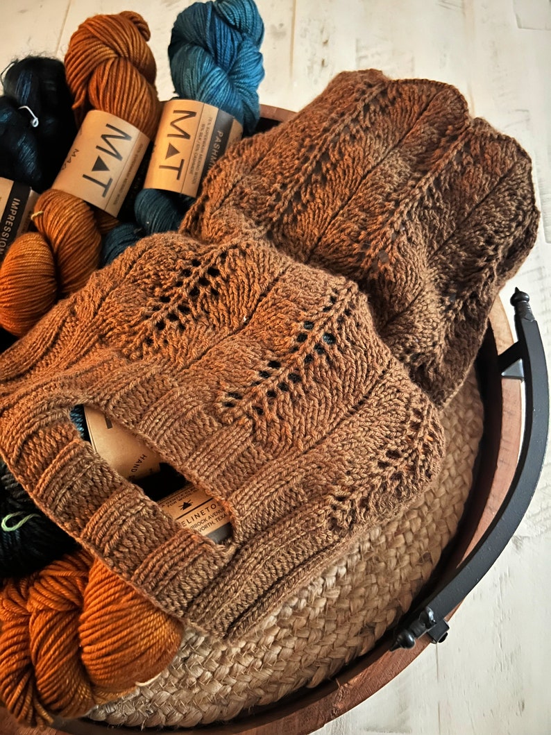 A rustic wooden basket filled with various colors of yarn and a knit market bag showcasing intricate patterns, placed on a white wooden surface, pattern available Marly Bird