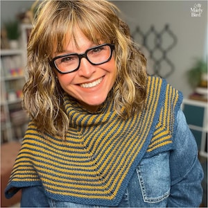 May include: A knitted cowl in shades of navy blue and mustard yellow, featuring a striped pattern. The cowl is worn over a denim jacket. The image also shows a person wearing black glasses and smiling.