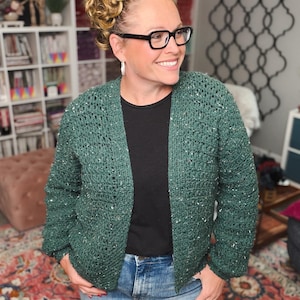 May include: A green crochet cardigan with white flecks. The open-front sweater is worn over a black top and blue jeans. The woman in the image is wearing black glasses and smiling.