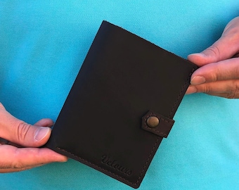 May include: Dark brown leather wallet with a snap closure. The wallet has a stitched border and the word "Velours" is embossed on the front. The wallet is held against a light blue background.