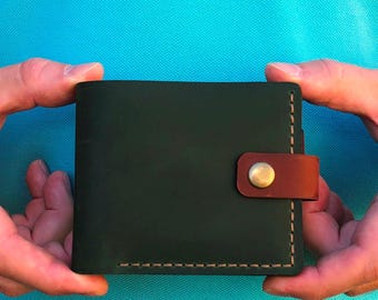 May include: Dark green leather wallet with a brown leather strap and a brass button closure. The wallet is square and has stitched edges. The background is a solid light blue.