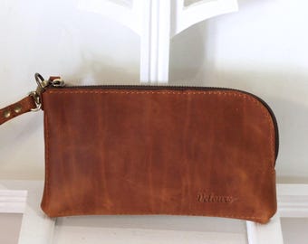 May include: A brown leather wristlet with a zipper closure and a wrist strap. The wristlet has a smooth texture and a small logo on the front. The wrist strap is attached with a metal clasp.