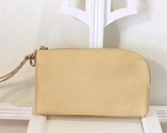 May include: A pale yellow leather wristlet pouch with a textured surface, a rounded right side, and a matching wrist strap attached to a silver-toned metal zipper closure. The pouch features a simple, rectangular shape with a curved corner.