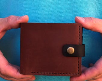 May include: A brown leather wallet with a black strap and a brass button closure. The wallet is square and has visible stitching around the edges. The wallet is held by two hands.