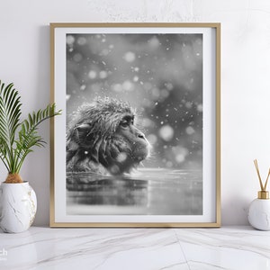 May include: Black and white framed print of a Japanese macaque, also known as a snow monkey, in a hot spring. The monkey is partially submerged in water, with snowflakes falling around it. The print is displayed on a white marble surface.