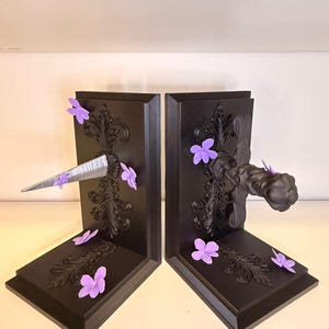 May include: A pair of black bookends with ornate scrollwork and purple flower accents. One bookend features a silver, metallic spike, while the other has a black, fist-shaped handle. Decorative and functional.