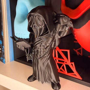 Könnte beinhalten: Eine schwarze Figur der Ghostface-Figur aus der Horrorfilmreihe Scream, die ein Buch hält. Die Figur steht auf einem weißen Regal, mit einem Buch und anderen dekorativen Elementen im Hintergrund.