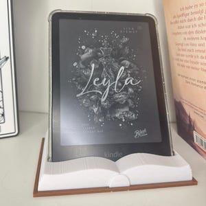 May include: A Kindle e-reader displaying the book "Leyla" by Lisa Kismet, held in a white book-shaped stand with a brown base. The e-reader has a clear protective case. The book cover features floral artwork.