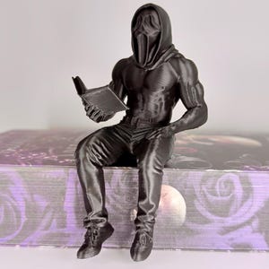 Könnte beinhalten: Eine schwarze 3D-gedruckte Figur einer muskulösen Figur mit Scream-Maske und Kapuzenpullover, die sitzt und ein Buch liest. Die Figur sitzt auf einem Buch mit floralem Design. Die Gesamtästhetik ist düster und gotisch.