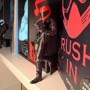 Peut inclure: Figurine noire d'une personne portant un casque de moto rouge et une veste, appuyée contre un mur et lisant un livre. Le mur porte le texte rouge "RUSH IN". La figurine est sur une étagère blanche.